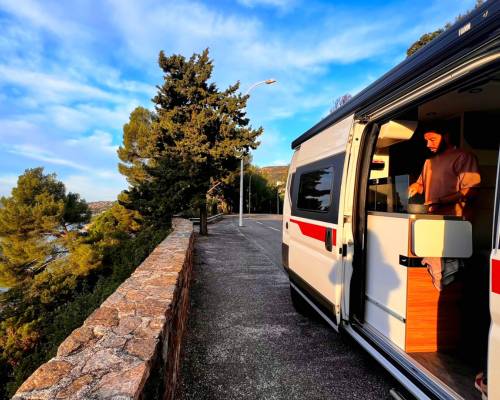 Man cooking inside an AVIS explore van, parked along a scenic tree-lined road overlooking the sea.