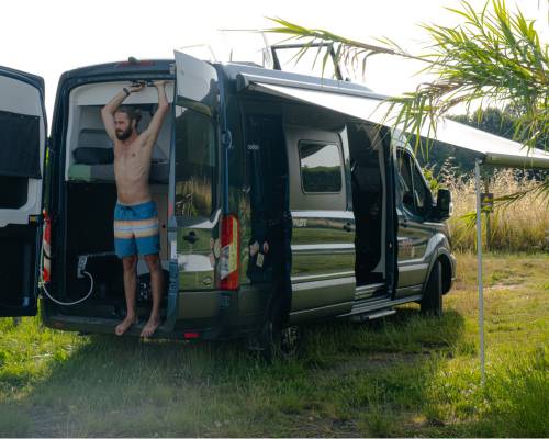 Man stretching out in the back of his AVIS van explores, with doors wide open to a verdant natural setting.