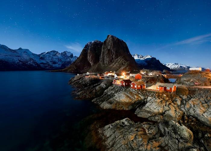 The fishing village of Hamnøy in the Lofoten Islands, Norway, with its traditional red houses nestled between snow-capped mountains and fjords, under a glittering sun and a striking sky.