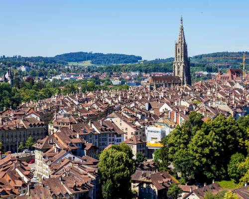 Panoramic view of the old town of Bern, the capital of Switzerland, with its historic red-roofed buildings lining the winding Aare River, surrounded by the green hills of the Bernese Pre-Alps